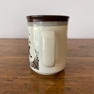 May include: A white ceramic mug with a brown rim and handle. The mug has a brown floral design on the side.