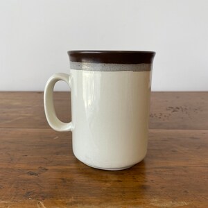 May include: A white ceramic mug with a brown rim and a brown band around the top. The mug is sitting on a wooden surface.