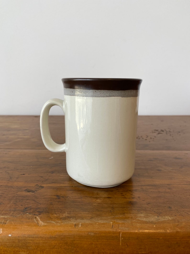 May include: A white ceramic mug with a brown rim and a brown band around the top. The mug is sitting on a wooden surface.