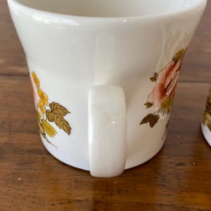 May include: White ceramic mug with a floral design featuring pink, yellow, and brown flowers. The mug has a white handle.