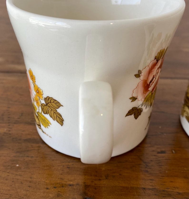 May include: White ceramic mug with a floral design featuring pink, yellow, and brown flowers. The mug has a white handle.