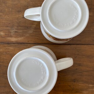 May include: Two white ceramic mugs with a floral design. The bottom of each mug reads "Made in New Zealand".