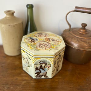 May include: A vintage, hexagonal tin featuring illustrations of women in hats and figures in period clothing. The tin is cream-coloured with gold trim and is on a wooden surface. Other vintage items are in the background.