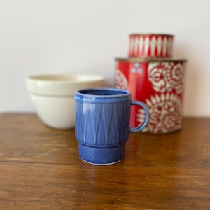May include: A blue ceramic mug with a textured, geometric design. The mug has a handle and sits on a wooden surface. In the background are a white bowl and red and white decorative containers.