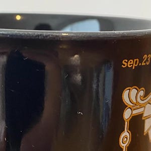 Pode incluir: Close-up de uma caneca de cer&acirc;mica preta com detalhes dourados. A caneca tem o texto "sep.23" em letras douradas. Um design decorativo dourado tamb&eacute;m &eacute; vis&iacute;vel.