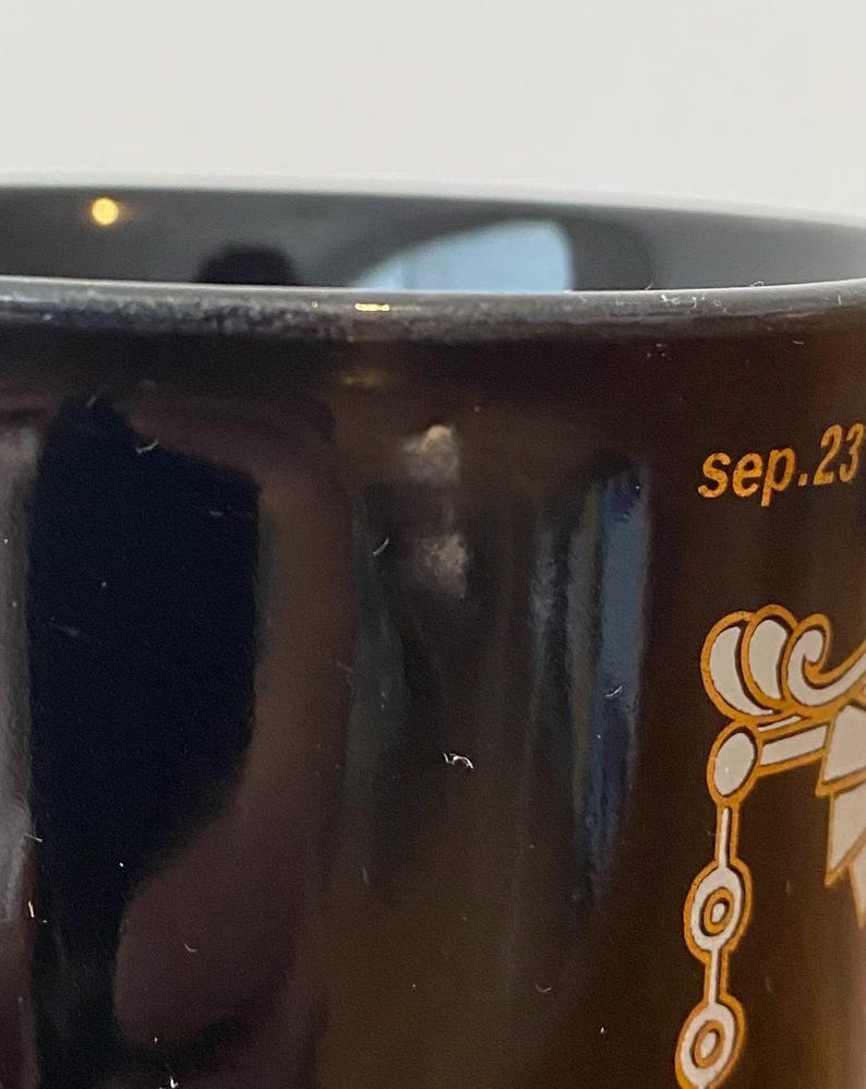 Pode incluir: Close-up de uma caneca de cer&acirc;mica preta com detalhes dourados. A caneca tem o texto "sep.23" em letras douradas. Um design decorativo dourado tamb&eacute;m &eacute; vis&iacute;vel.
