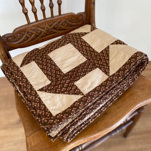 May include: A folded quilt featuring a geometric pattern of solid beige squares and dark brown fabric printed with small tan cross and circle motifs. The quilt is constructed with stitched seams and is layered in a stack on a wooden chair.