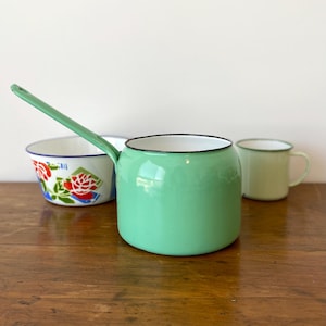 May include: A selection of vintage enamelware kitchen items. A mint green saucepan with a long handle, a white bowl with a floral design, and a light green mug with a black rim are arranged on a wooden surface.