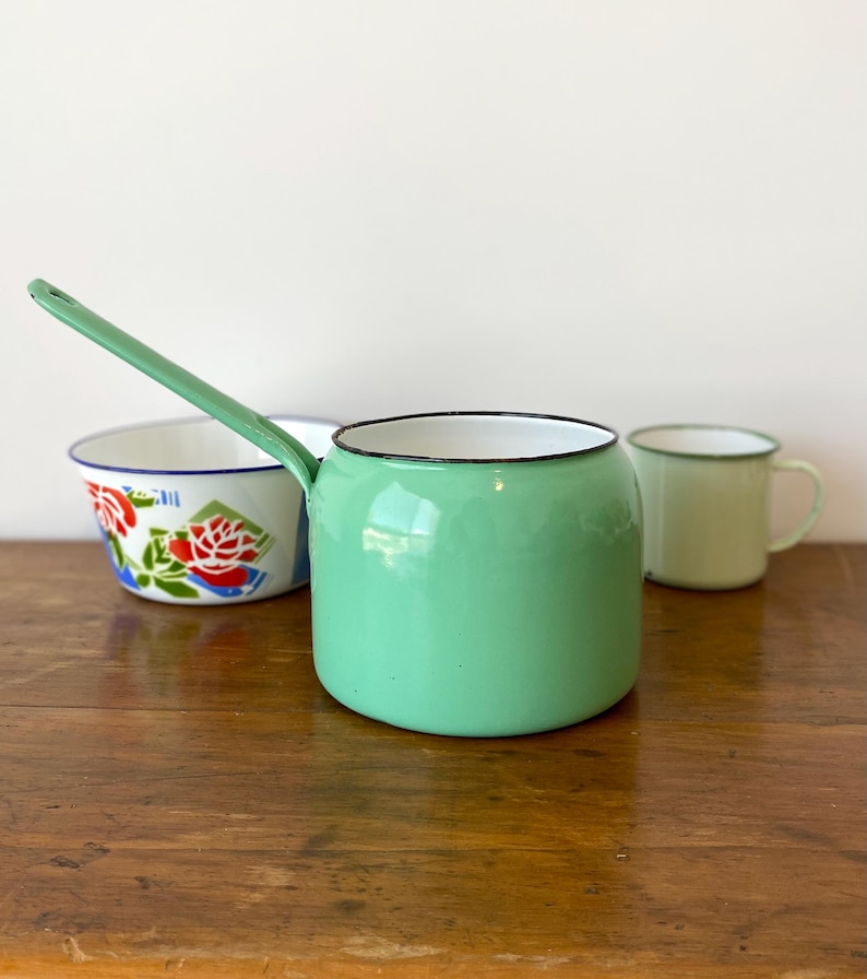 May include: A collection of vintage enamelware kitchen items. A mint green saucepan with a long handle, a white bowl with a floral design, and a light green mug with a black rim are arranged on a wooden surface.