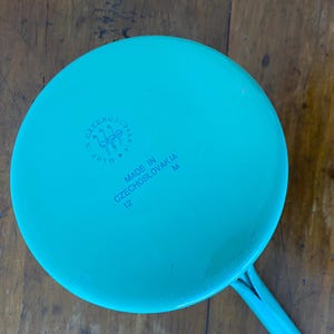 May include: A teal-colored saucepan with a long handle. The bottom of the pan features the text "MADE IN CZECHOSLOVAKIA" and the number "12". A circular emblem is also visible.