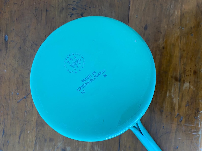 May include: A teal-colored saucepan with a long handle. The bottom of the pan features the text "MADE IN CZECHOSLOVAKIA" and the number "12". A circular emblem is also visible.