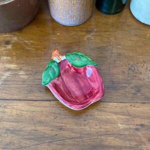 Pode incluir: Um prato de cerâmica em forma de maçã com corpo vermelho escuro, folhas verdes e borda branca. O prato é colocado sobre uma superfície de madeira, com outros objetos de cerâmica ao fundo.