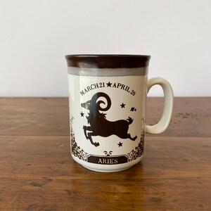 May include: A white ceramic mug with a brown rim and handle. The mug features a brown silhouette of an Aries zodiac symbol with the text "MARCH 21 * APRIL 20" and "ARIES" below the symbol.