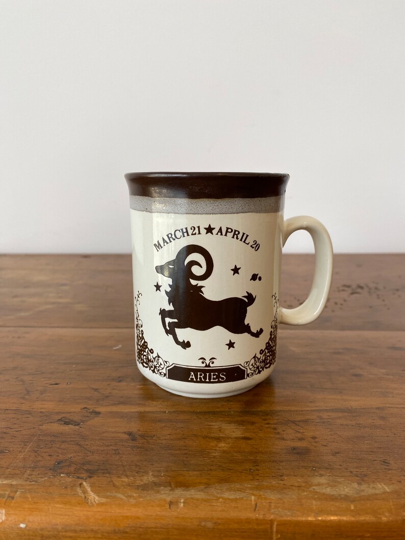 May include: A white ceramic mug with a brown rim and handle. The mug features a brown silhouette of an Aries zodiac symbol with the text "MARCH 21 * APRIL 20" and "ARIES" below the symbol.
