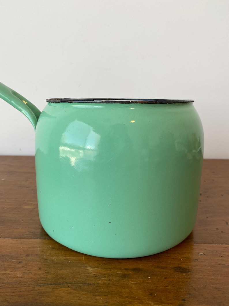 May include: A mint green enamel saucepan with a long handle. The pot has a rounded body and a black rim. The handle is also mint green. The saucepan is sitting on a wooden surface.