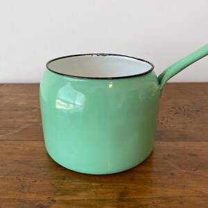 May include: A vintage, mint green enamel saucepan with a long handle. The pot has a white interior and a black rim. The saucepan is sitting on a wooden surface.