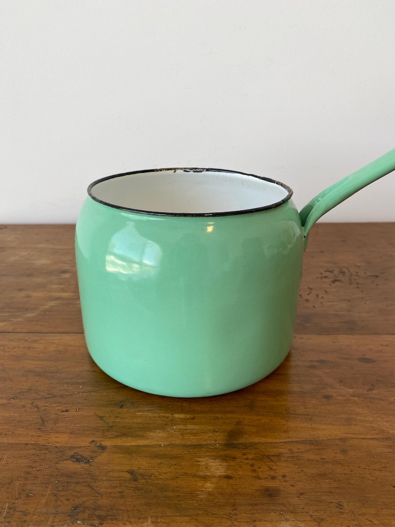 May include: A vintage, mint green enamel saucepan with a long handle. The pot has a white interior and a black rim. The saucepan is sitting on a wooden surface.