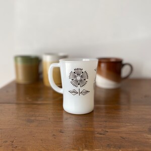 May include: White ceramic mug with a black geometric flower design. The mug has a handle and is sitting on a wooden table.