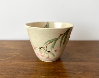 May include: A cream-colored ceramic bowl with a tapered shape, featuring a hand-painted design of green leaves and small pink flowers on the exterior. The bowl has a smooth, matte finish and a wide, open rim, showcasing a simple, functional design.