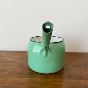 May include: A mint green enamelware kettle with a black rim and handle. The kettle has a rounded body and a spout with a hole. The handle is attached to the side of the kettle. The kettle is sitting on a wooden surface.