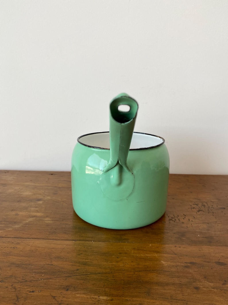 May include: A mint green enamelware kettle with a black rim and handle. The kettle has a rounded body and a spout with a hole. The handle is attached to the side of the kettle. The kettle is sitting on a wooden surface.