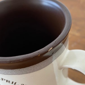 May include: A white ceramic mug with a brown rim and a brown band around the middle. The mug has a crack in the rim and the text "APRIL 20" printed on the front.