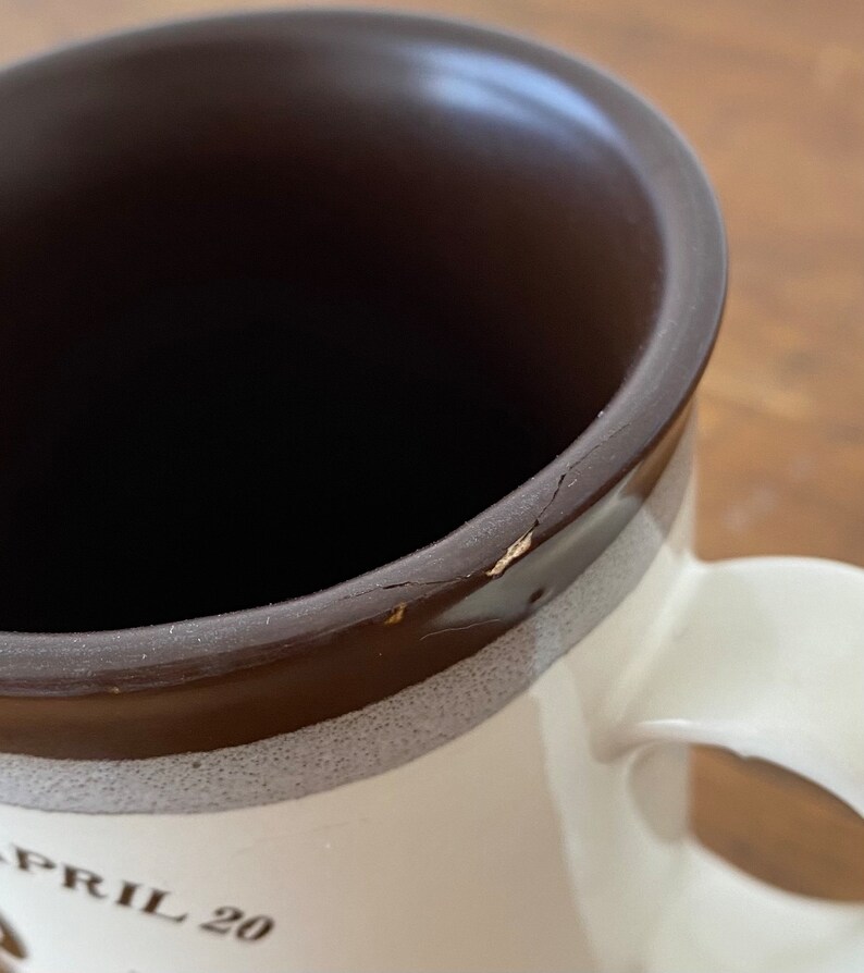 May include: A white ceramic mug with a brown rim and a brown band around the middle. The mug has a crack in the rim and the text "APRIL 20" printed on the front.
