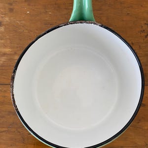 May include: A vintage white enamel saucepan with a green handle. The rim is outlined in black, with a distressed brown edge. The handle is a solid, curved design. The pan sits on a wooden surface.