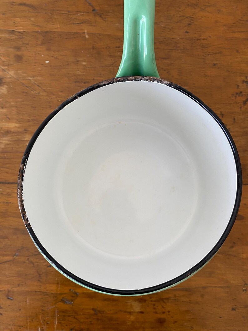 May include: A vintage white enamel saucepan with a green handle. The rim is outlined in black, with a distressed brown edge. The handle is a solid, curved design. The pan sits on a wooden surface.