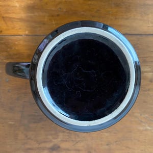 Pode incluir: Uma caneca de cer&acirc;mica preta com uma borda branca, vista de baixo. A caneca tem um acabamento brilhante e uma al&ccedil;a. O interior da caneca &eacute; escuro e a caneca est&aacute; em uma superf&iacute;cie de madeira.