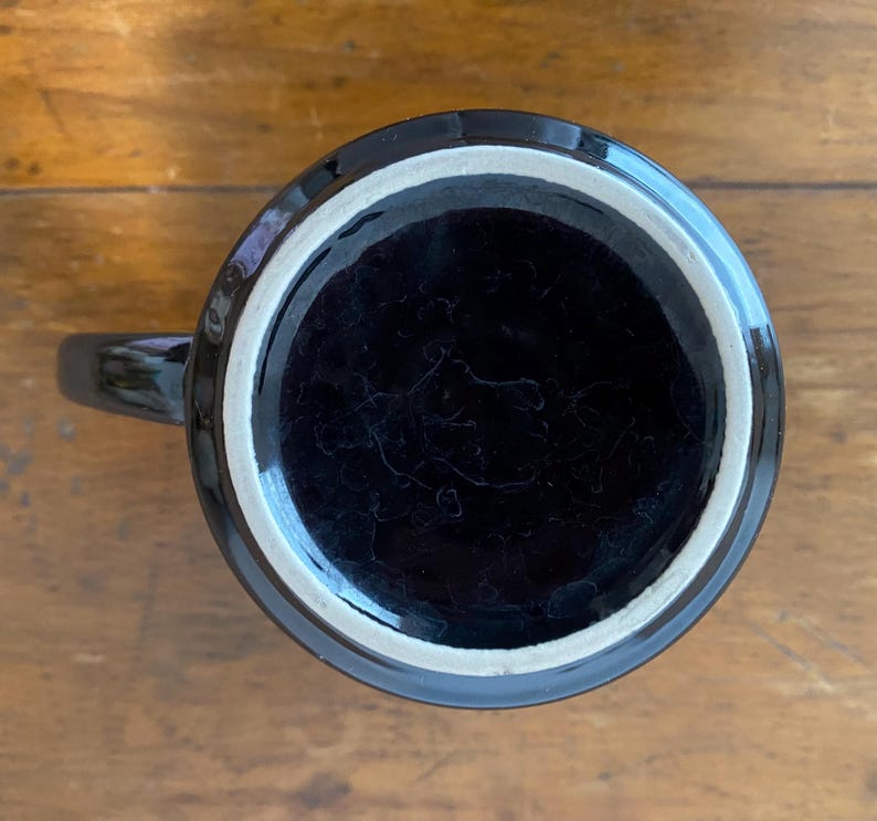 Pode incluir: Uma caneca de cer&acirc;mica preta com uma borda branca, vista de baixo. A caneca tem um acabamento brilhante e uma al&ccedil;a. O interior da caneca &eacute; escuro e a caneca est&aacute; em uma superf&iacute;cie de madeira.