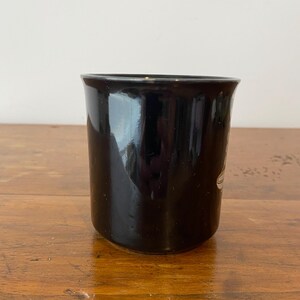 Pode incluir: Uma caneca de cer&acirc;mica preta com acabamento brilhante. A caneca tem formato cil&iacute;ndrico com uma borda ligeiramente alargada. Um desenho branco &eacute; vis&iacute;vel na lateral da caneca. A caneca est&aacute; sobre uma superf&iacute;cie de madeira.