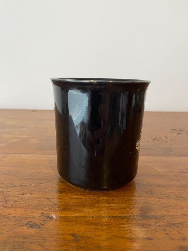 Pode incluir: Uma caneca de cer&acirc;mica preta com acabamento brilhante. A caneca tem formato cil&iacute;ndrico com uma borda ligeiramente alargada. Um desenho branco &eacute; vis&iacute;vel na lateral da caneca. A caneca est&aacute; sobre uma superf&iacute;cie de madeira.