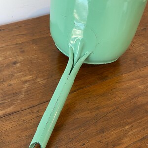 May include: A vintage, mint green dustpan with a long handle. The dustpan is made of metal and has a rounded shape with a flat bottom. The handle has a hole at the end for hanging. The dustpan is resting on a wooden surface.