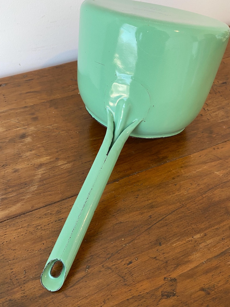 May include: A vintage, mint green dustpan with a long handle. The dustpan is made of metal and has a rounded shape with a flat bottom. The handle has a hole at the end for hanging. The dustpan is resting on a wooden surface.