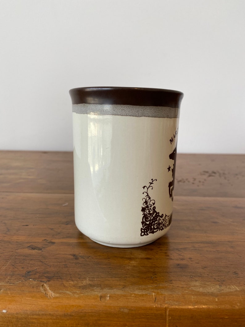 May include: A white ceramic mug with a brown rim and a brown design of a horse and floral pattern. The mug says "Magic" on the side.