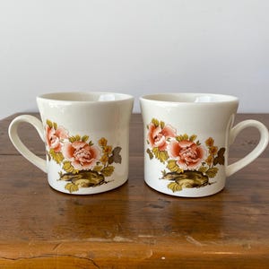 May include: Two white ceramic mugs with floral designs. The mugs have a pink and yellow floral pattern with green leaves. The mugs have a handle on the side.