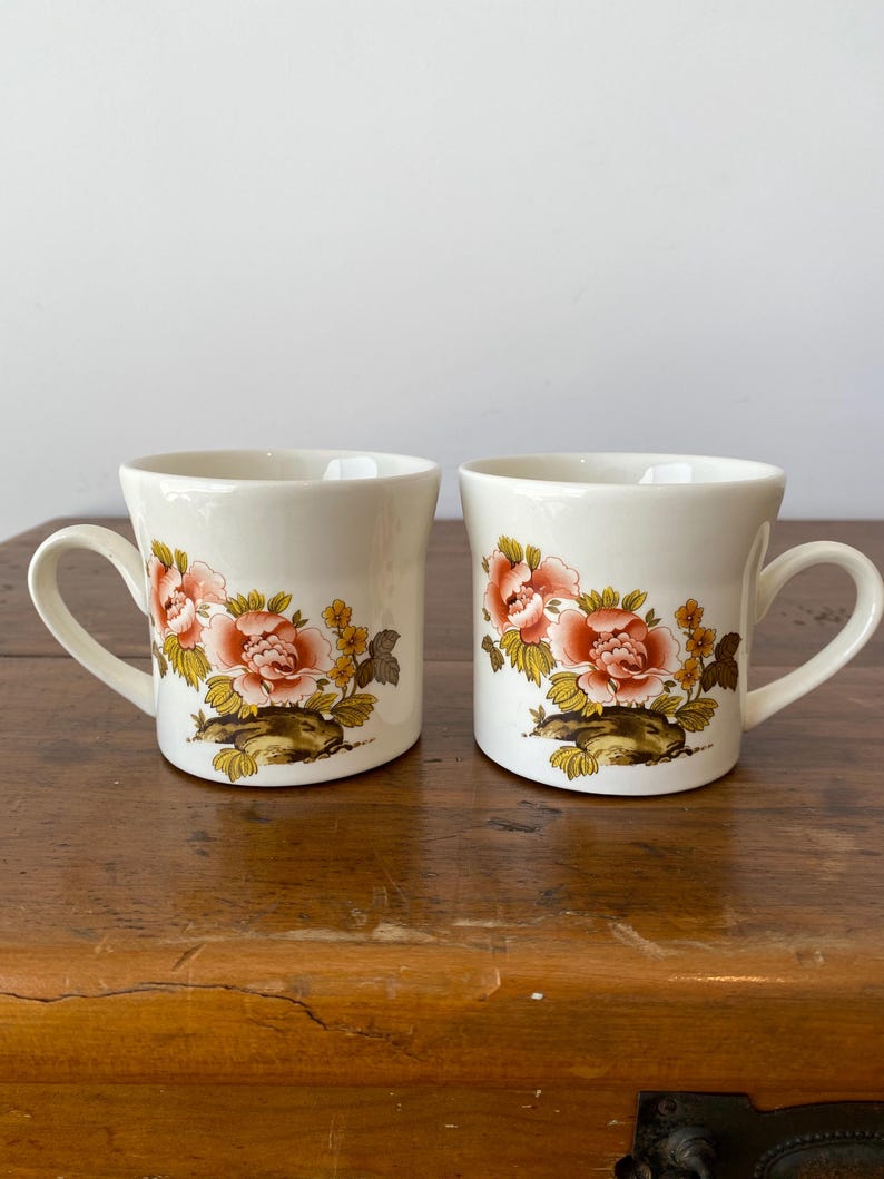 May include: Two white ceramic mugs with floral designs. The mugs have a pink and yellow floral pattern with green leaves. The mugs have a handle on the side.