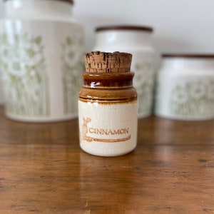 May include: A small white ceramic jar with a brown lid and cork stopper. The jar is labelled "CINNAMON" in brown lettering. The jar is sitting on a wooden surface.