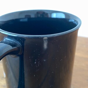 Pode incluir: Um close-up de uma caneca de caf&eacute; preta com uma al&ccedil;a curva. A caneca tem um acabamento brilhante e parece estar vazia. O interior da caneca &eacute; escuro e a borda &eacute; lisa. A caneca est&aacute; em uma superf&iacute;cie de madeira.