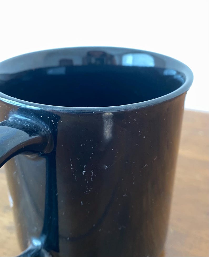Pode incluir: Um close-up de uma caneca de caf&eacute; preta com uma al&ccedil;a curva. A caneca tem um acabamento brilhante e parece estar vazia. O interior da caneca &eacute; escuro e a borda &eacute; lisa. A caneca est&aacute; em uma superf&iacute;cie de madeira.