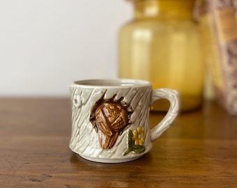 May include: A ceramic mug with a tree bark design and a brown owl emerging from a hole. The mug has a small yellow flower and a textured handle. The mug is on a wooden surface.