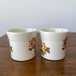 May include: Two white ceramic mugs with floral designs. The mugs have a rounded shape and a floral pattern in shades of pink, yellow, and green.