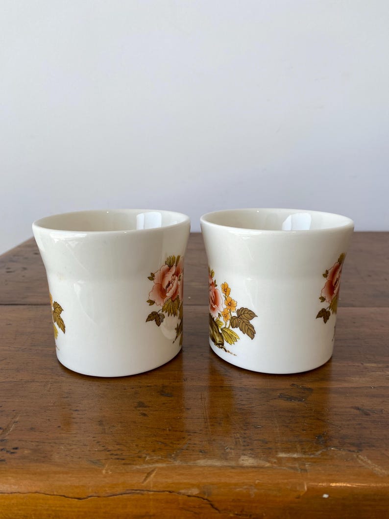 May include: Two white ceramic mugs with floral designs. The mugs have a rounded shape and a floral pattern in shades of pink, yellow, and green.