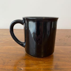 Pode incluir: Uma caneca de cer&acirc;mica preta brilhante com uma al&ccedil;a curva. A caneca tem uma forma simples e cil&iacute;ndrica e uma superf&iacute;cie lisa e reflexiva. A caneca est&aacute; em uma superf&iacute;cie de madeira.