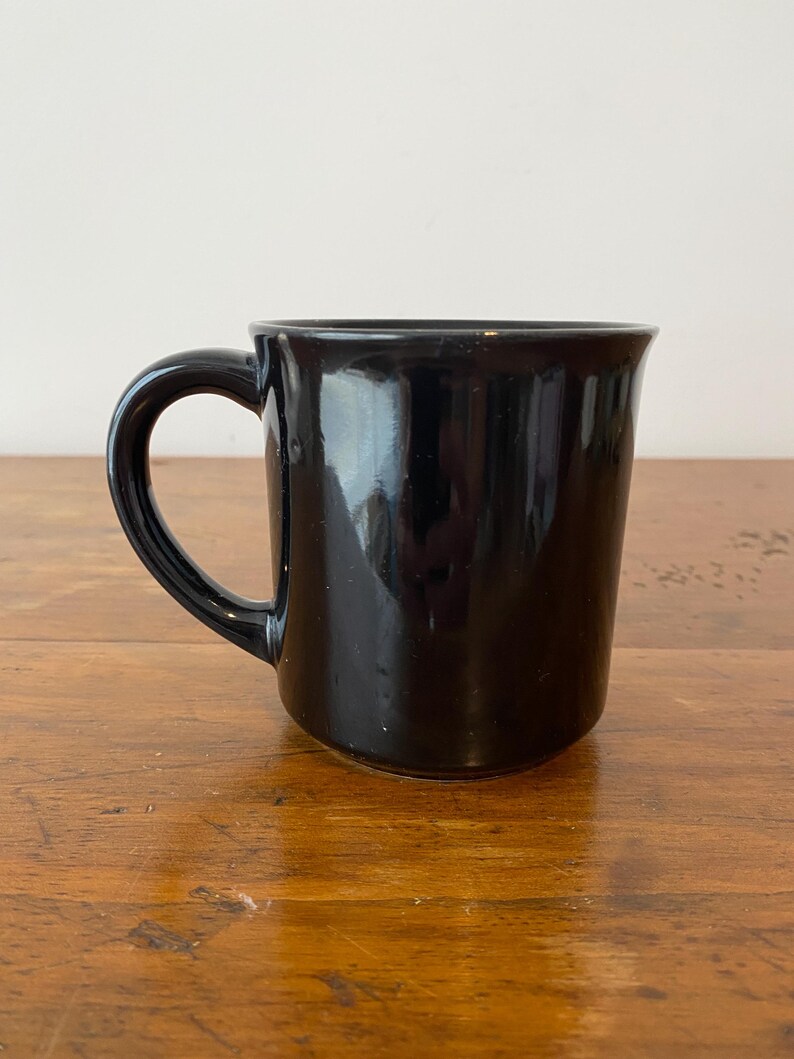 Pode incluir: Uma caneca de cer&acirc;mica preta brilhante com uma al&ccedil;a curva. A caneca tem uma forma simples e cil&iacute;ndrica e uma superf&iacute;cie lisa e reflexiva. A caneca est&aacute; em uma superf&iacute;cie de madeira.