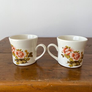 May include: Two white ceramic mugs with floral designs. The mugs have a pink, yellow, and brown floral pattern. The mugs are on a wooden surface.