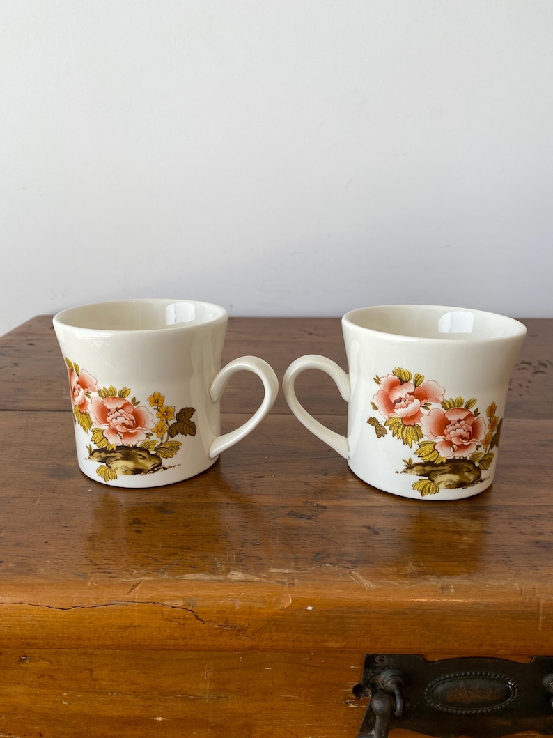 May include: Two white ceramic mugs with floral designs. The mugs have a pink, yellow, and brown floral pattern. The mugs are on a wooden surface.