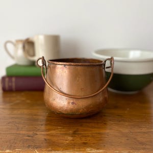 Pode incluir: Uma pequena panela de cobre arredondada com um acabamento metálico desgastado e uma alça de cobre fina e arqueada presa a dois anéis laterais. A panela apresenta uma abertura larga e um corpo curvo com sinais visíveis de envelhecimento.