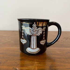 Pode incluir: Caneca de cer&acirc;mica preta com um design do signo do zod&iacute;aco de Libra em dourado e branco. A caneca tem uma al&ccedil;a e a palavra "LIBRA" em letras douradas. As datas "sep.23 - oct.23" tamb&eacute;m est&atilde;o impressas na caneca.